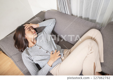 Indonesian southeast asian woman with abdominal pain due to constipation or menstruation issues. Suffering from stomach aches, cramps, and holding her belly on a couch at home. Healthcare concept Indonesian southeast asian woman with abdominal pain due to constipation or menstruation issues. Suffering from stomach aches, cramps, and holding her belly on a couch at home. Healthcare concept 127862782
