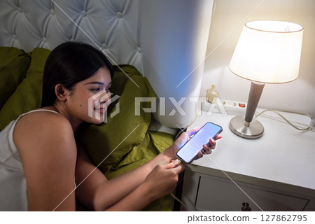 Indonesian southeast asian woman lying down on a bed and charging her cellphone at night before sleep at home Indonesian southeast asian woman lying down on a bed and charging her cellphone at night before sleep at home 127862795