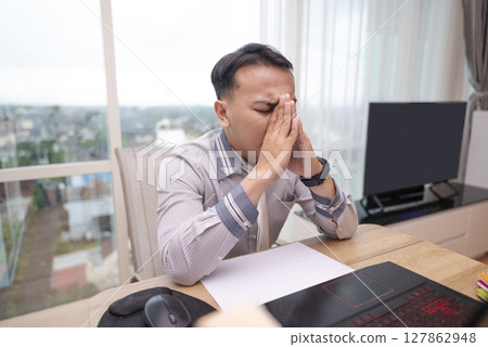 Indonesian southeast asian businessman experiencing stress and burnout while working on laptop in hybrid workplace. Paperwork and a cellphone on the table. Business concept. Remote working from home 127862948