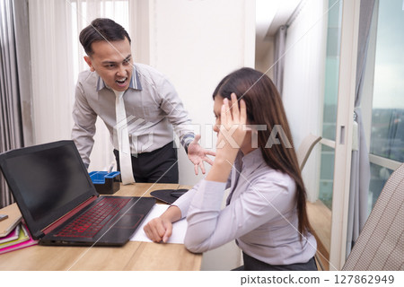 Indonesian southeast asian businessman scolding his frustrated employee for being late, pointing at his watch, the stress of deadlines. Laptop and paperwork on the table. Business concept 127862949