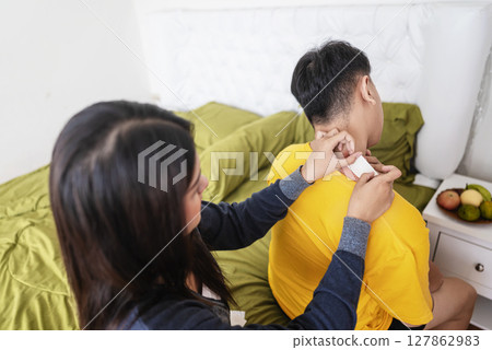 Indonesian southeast asian woman applies a neck pain patch to her husband neck in the bedroom. Self-care and wellness routines concept 127862983