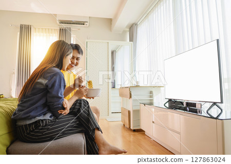 Indonesian southeast asian couple family eating noodles meal together while watching television at home. Empty television screen for mockup, template, or copy space Indonesian southeast asian couple family eating noodles meal together while watching television at home. Empty television screen for mockup, template, or copy space 127863024