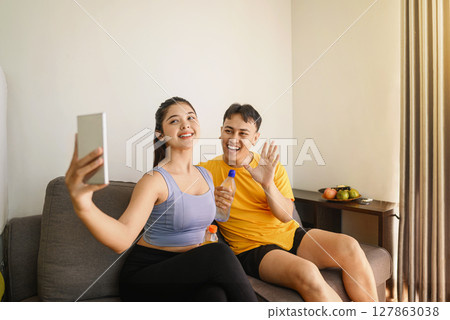 Indonesian southeast asian couple drinking water while taking a selfie or video call with their cellphone after finishing a home body workout exercise session. Fitness and active lifestyles concept 127863038