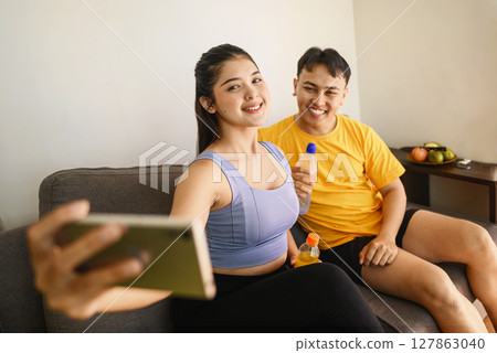Indonesian southeast asian couple drinking water while taking a selfie or video call with their cellphone after finishing a home body workout exercise session. Fitness and active lifestyles concept Indonesian southeast asian couple drinking water while taking a selfie or video call with their cellphone after finishing a home body workout exercise session. Fitness and active lifestyles concept 127863040
