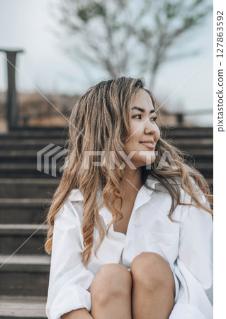 Asian young woman with wavy hair on wooden stairs in white shirt, with hand in hair, peaceful emotional moment outdoors, mindfulness, feminine wellness, emotional balance 127863592