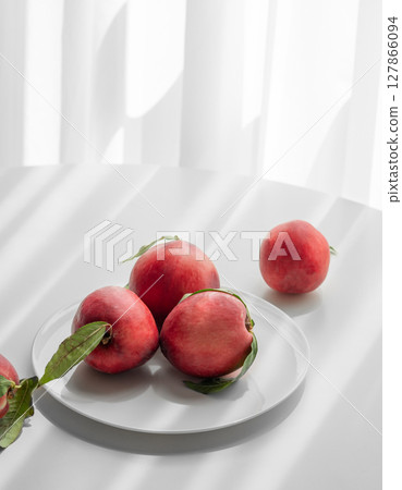 Fresh red peaches or nectarines with green leaves, on plate on white table and sunlight from window. 127866094