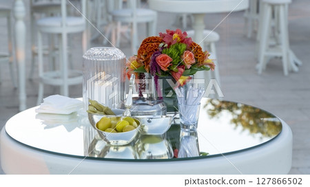 An elegant table setting with a floral centerpiece, olives, pickles, and drink stirrers on a reflective surface, with blurred chairs and tables in background 127866502