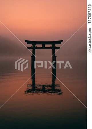 A torii gate on the lake surface dyed in the morning glow, a mysterious and tranquil Japanese landscape shrouded in mist 127867336