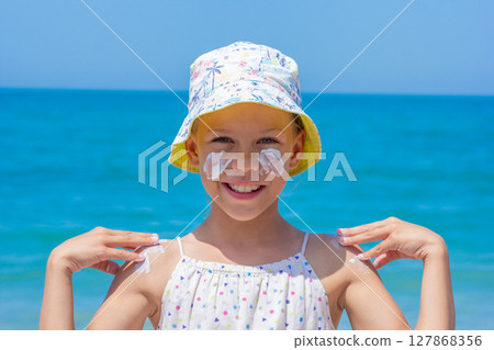 Child happily applying sunscreen on her face and shoulders at the beach, wearing a vibrant hat Child happily applying sunscreen on her face and shoulders at the beach, wearing a vibrant hat 127868356