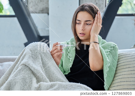 Sad upset teenage girl looking at thermometer sitting at home under blanket 127869803