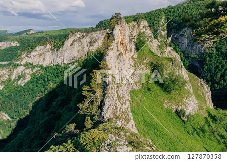 Dramatic rocky cliff surrounded by lush forest and green hills under a summer sky, captured in daylight. 127870358