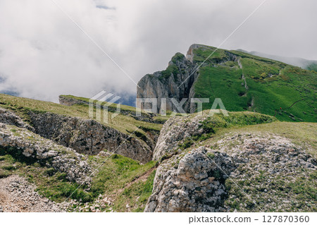 Mountain ridge with green slopes and dramatic cliffs rising above clouds on a moody summer day. Bolshoy Tkhach national park Mountain ridge with green slopes and dramatic cliffs rising above clouds on a moody summer day. Bolshoy Tkhach national park 127870360