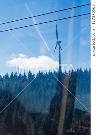 A towering wind turbine rises confidently against a tranquil blue sky, casting its gaze over the vibrant green forests below on a brilliant afternoon A towering wind turbine rises confidently against a tranquil blue sky, casting its gaze over the vibrant green forests below on a brilliant afternoon 127870569