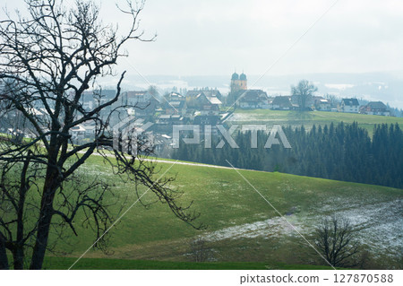 Breathtaking view of a serene village nestled in rolling green hills under a cloudy sky in early springtime, capturing the essence of tranquility and nature's beauty 127870588