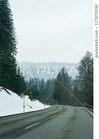 A meandering road stretches through a picturesque snowy landscape, adorned with stately evergreen trees, all beneath a serene, cloudy sky that enhances the tranquil beauty of the winter scenery 127870590