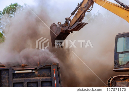 Heavy machinery operates to load construction debris into truck, creating cloud of dust at works site. 127871170