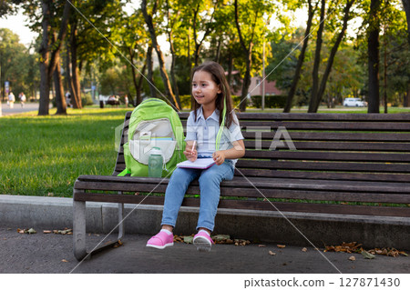 Young girl enjoying outdoor park with notepad and backpack on bench 127871430