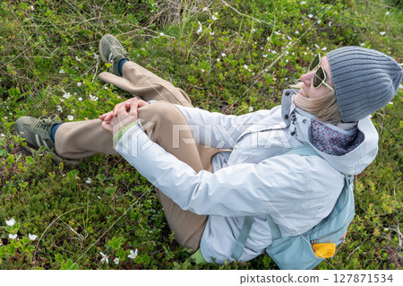 Smiling woman in a gray beanie, white jacket, and sunglasses relaxing on green tundra among blooming cloudberry flowers in Teriberka Nature Reserve 127871534