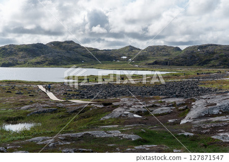 Scenic wooden trail winding through a remote Arctic landscape with tundra, rocky hills, and a lake under dramatic clouds 127871547