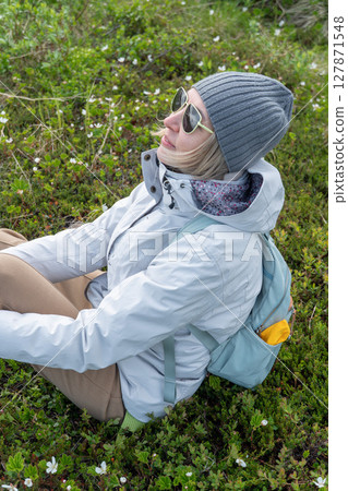 Smiling woman in a gray beanie, white jacket, and sunglasses relaxing on green tundra among blooming cloudberry flowers in Teriberka Nature Reserve Smiling woman in a gray beanie, white jacket, and sunglasses relaxing on green tundra among blooming cloudberry flowers in Teriberka Nature Reserve 127871548