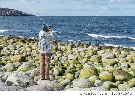 Woman in light hiking outfit takes a photo of the Barents Sea on Dragon Eggs Beach in Teriberka, standing among round moss-covered boulders Woman in light hiking outfit takes a photo of the Barents Sea on Dragon Eggs Beach in Teriberka, standing among round moss-covered boulders 127871549