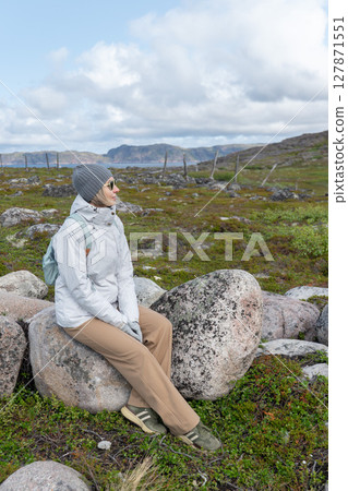 Smiling woman in a white jacket sitting on a large stone surrounded by tundra landscape, with mountains and cloudy sky in the background Smiling woman in a white jacket sitting on a large stone surrounded by tundra landscape, with mountains and cloudy sky in the background 127871551