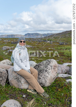Smiling woman in a white jacket sitting on a large stone surrounded by tundra landscape, with mountains and cloudy sky in the background 127871552