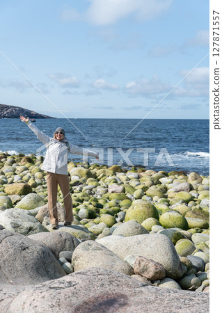 Woman in light hiking clothes with outstretched arms stands joyfully on the rocky Dragon Eggs Beach in Teriberka, Russia, facing the Barents Sea 127871557