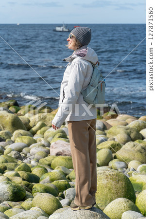 Woman stands on the unique round stones at the Dragon Eggs Beach in Teriberka, looking at the Barents Sea with a peaceful expression 127871560