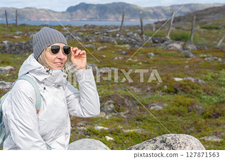 Woman in a white jacket adjusting her sunglasses while sitting among tundra rocks in Teriberka Nature Reserve with mountains and sea 127871563