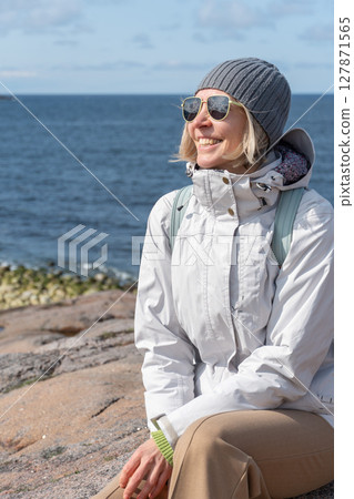 Smiling woman in a gray beanie and white jacket sits on a rocky coastline with the sea in the background under a blue sky 127871565