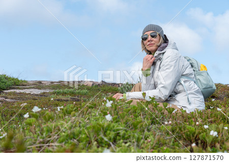 Woman in a gray beanie, white jacket, and sunglasses sitting among arctic vegetation in Teriberka Nature Reserve under a clear blue sky 127871570