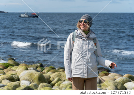Woman smiles while standing on the distinctive rounded boulders at Dragon Eggs Beach in Teriberka, with the Barents Sea and a boat in the background Woman smiles while standing on the distinctive rounded boulders at Dragon Eggs Beach in Teriberka, with the Barents Sea and a boat in the background 127871583