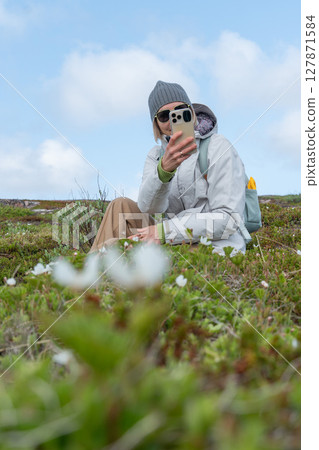 Woman in a gray beanie and sunglasses taking a selfie while sitting among wild Arctic flowers in Teriberka Nature Reserve under a blue sky Woman in a gray beanie and sunglasses taking a selfie while sitting among wild Arctic flowers in Teriberka Nature Reserve under a blue sky 127871584