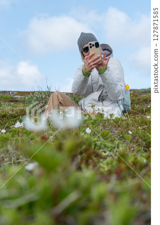 Woman in a gray beanie and sunglasses taking a selfie while sitting among wild Arctic flowers in Teriberka Nature Reserve under a blue sky Woman in a gray beanie and sunglasses taking a selfie while sitting among wild Arctic flowers in Teriberka Nature Reserve under a blue sky 127871585