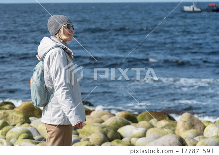 Woman in a light jacket and grey beanie stands calmly on the moss-covered boulders of Dragon Eggs Beach in Teriberka, looking at the Barents Sea 127871591