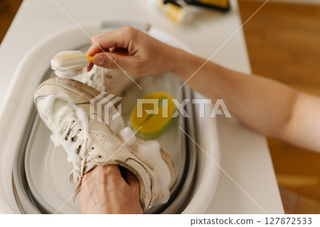 Close-up top view of male hands scrubbing muddy white sneakers with small brush and soap in white basin at home. Concept of shoe care, maintenance and cleanliness. 127872533
