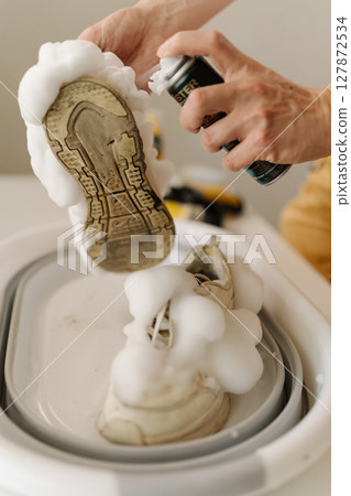 Close-up of young man applying foam cleaner on dirty white sneakers in basin, highlighting shoe cleaning process and emphasizing proper care. Concept of shoe maintenance and hygiene. Close-up of young man applying foam cleaner on dirty white sneakers in basin, highlighting shoe cleaning process and emphasizing proper care. Concept of shoe maintenance and hygiene. 127872534