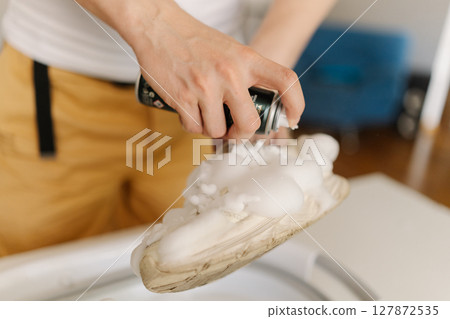 Close-up cropped shot of young man spraying foam cleaner on dirty white shoe, holding above washing basin, ready for thorough cleaning at home. Concept of shoe maintenance and hygiene. Close-up cropped shot of young man spraying foam cleaner on dirty white shoe, holding above washing basin, ready for thorough cleaning at home. Concept of shoe maintenance and hygiene. 127872535