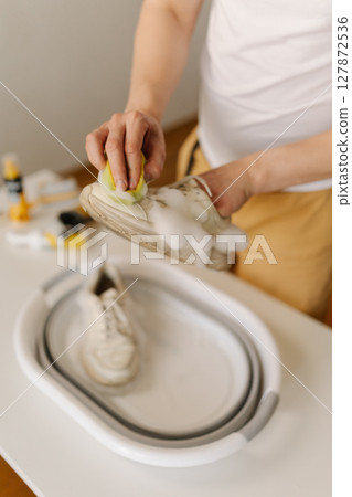Close-up vertical shot of unrecognizable housekeeper washing dirty white sneakers using small yellow sponge and soap in white foldable basin on table, with cleaning products on blurred background. Close-up vertical shot of unrecognizable housekeeper washing dirty white sneakers using small yellow sponge and soap in white foldable basin on table, with cleaning products on blurred background. 127872536