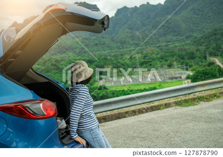 Asian woman enjoying peaceful mountain vista during sustainable road trip adventure with open SUV car tailgate on eco friendly summer vacation exploring green forest landscape for wellness retreat. Asian woman enjoying peaceful mountain vista during sustainable road trip adventure with open SUV car tailgate on eco friendly summer vacation exploring green forest landscape for wellness retreat. 127878790