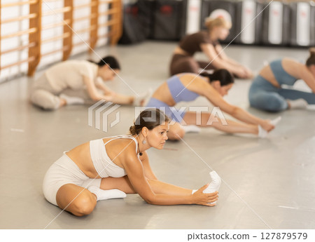 Group of girls in gym do stretching, sitting on floor and bending towards legs. Group of girls in gym do stretching, sitting on floor and bending towards legs. 127879579