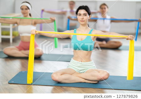 Group of women sitting on sports mats and doing arm stretches using resistance bands at Pilates class Group of women sitting on sports mats and doing arm stretches using resistance bands at Pilates class 127879826