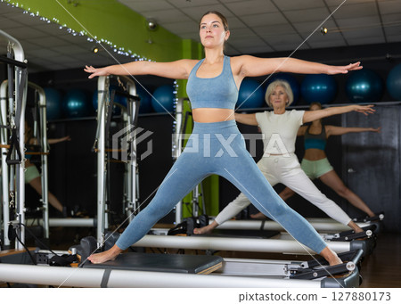 Young woman standing on pilates reformer, perform exercises Young woman standing on pilates reformer, perform exercises 127880173