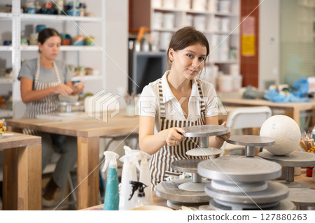 Girl in pottery workshop stands at table with tools, picks up rotate turntable revolving for work 127880263