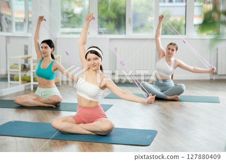 Girl conducts Pilates class for girls sitting with elastic band in hands above head makes body bends 127880409