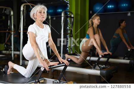 Senior woman standing on knee reformer, perform exercises and body tilts. 127880558