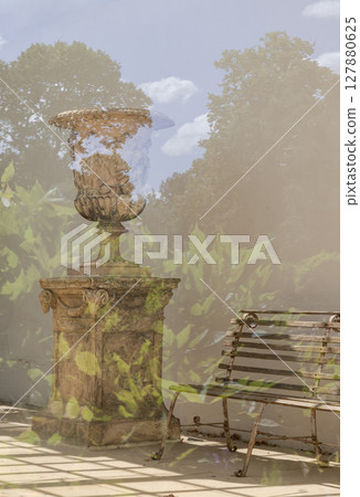 A decorative vintage stone urn and pedestal positioned next to a weathered wooden bench, with natural light. 127880625