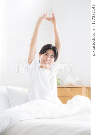 一位年輕人在床上醒來，沐浴在早晨的陽光下伸展雙臂 127882148