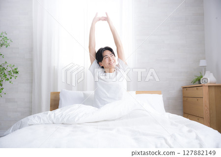 一位年輕人在床上醒來,沐浴在早晨的陽光下伸展雙臂 一位年輕人在床上醒來,沐浴在早晨的陽光下伸展雙臂 127882149